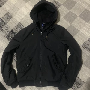 Svart jacka från H&M Divided - Säljer en svart jacka från H&M Divided med huva och dragkedja. Jackan har ribbade muddar och två fickor med knappar framtill. Perfekt för en casual look.