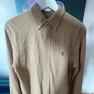 Ralph Lauren skjorta  - Snygg beige skjorta från Ralph Lauren med klassisk krage och knappar framtill. Skjortan har en broderad logotyp på bröstet och är långärmad. Perfekt för en stilren look i storlek M. Skick 9/10✅ Skriv vid frågor eller funderingar i DM🍾