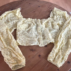 Gul blommig blus - Charmig gul blus med blommigt mönster och långa ärmar. Blusen har elastiska ärmslut och en luftig passform, 