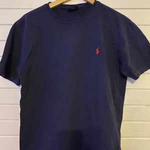 Mörkblå t-shirt från Polo Ralph Lauren - Säljer en klassisk mörkblå t-shirt från Polo Ralph Lauren med det ikoniska röda broderade logotypen på bröstet. Perfekt för en stilren och avslappnad look. 👕