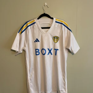 Äkta Leeds United fotbollströja från Adidas - Säljer en vit fotbollströja från Adidas med korta ärmar. Tröjan har blå och gula detaljer på axlarna och ett klubbmärke på bröstet. Perfekt för fotbollsfans som vill visa sitt stöd.