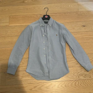 Blå skjorta från Polo Ralph Lauren - Snygg blå skjorta från Polo Ralph Lauren med klassisk krage och knappar framtill.slimfit. Perfekt för en stilren look. Den har en broderad logotyp på bröstet och är långärmad.tveka inte att skriva om du har någon fråga.pris kan diskuteras vid snabb affär 