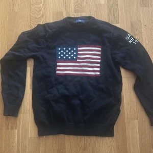 Svart tröja med amerikansk flagga från GANT - Säljer en svart tröja från GANT med en stor amerikansk flagga på framsidan och texten 'GANT NH-CT 1949' på ärmen. Tröjan har långa ärmar och en rund halsringning. Perfekt för en stilren och avslappnad look.