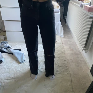 Svarta jeans med slitningar - Snygga svarta jeans med hög midja och slitningar vid knäna. De har en straight passform och är perfekta för en trendig look. Jeansen är tillverkade i ett robust denimtyg.
