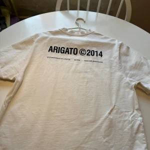 Vit t-shirt från Axel Arigato - Säljer en stilren vit t-shirt från Axel Arigato med tryck 'ARIGATO ©2014' på bröstet och ryggen. Perfekt för en skön vardagslook