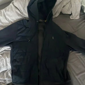 Svart cardigan från Ralph Lauren - Säljer en stilren svart cardigan från Ralph Lauren med dragkedja och huva. Jackan har ett diskret broderat logotyp på bröstet och är perfekt för kyligare dagar. Den har långa ärmar och en quiltad design som ger extra värme.