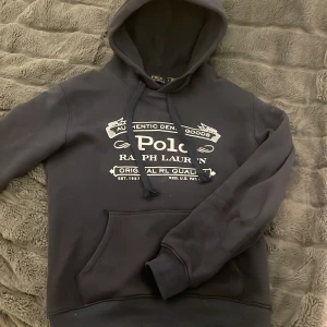 Blå hoodie från Ralph Lauren - Säljer en blå hoodie från Ralph Lauren med tryck på framsidan. Hoodien har en klassisk design med dragsko i huvan och en stor magficka. Perfekt för en avslappnad stil.