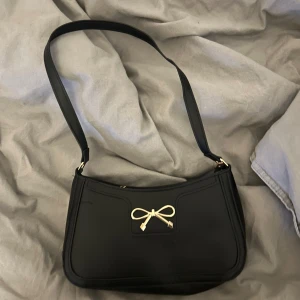 Svart axelväska med rosett - Snygg svart axelväska med en elegant rosett-detalj i guld på framsidan. Väskan har en enkel och stilren design med en justerbar axelrem. Perfekt för att ge en touch av elegans till din outfit.