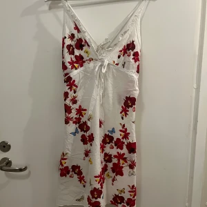 Vit blommig klänning från Sand&Sun - Säljer en somrig vit klänning från Sand&sun med röda blommor och fjärilar. Klänningen har en v-ringad design och justerbar knytning framtill. Perfekt för varma beachdagar. Det står att det är storlek XL men passar bra på mig som är storlek S