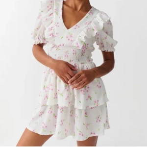 Blommig Gina klänning - Charmig vit klänning med blommigt mönster i rosa och grönt. Den har volanger vid ärmar och hals samt en v-ringad design. Perfekt för sommarens festligheter. Använd endast 1 gång och det finns inga täcken på användning. Skriv för fler bilder💕