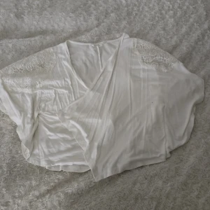 Vit omlottopp från KappAhl - Säljer en vit omlottopp från KappAhl med vackra spetsdetaljer på axlarna. Toppen har en lös passform och korta ärmar, perfekt för en stilren look. Passar bra till både jeans och kjol. 🌟