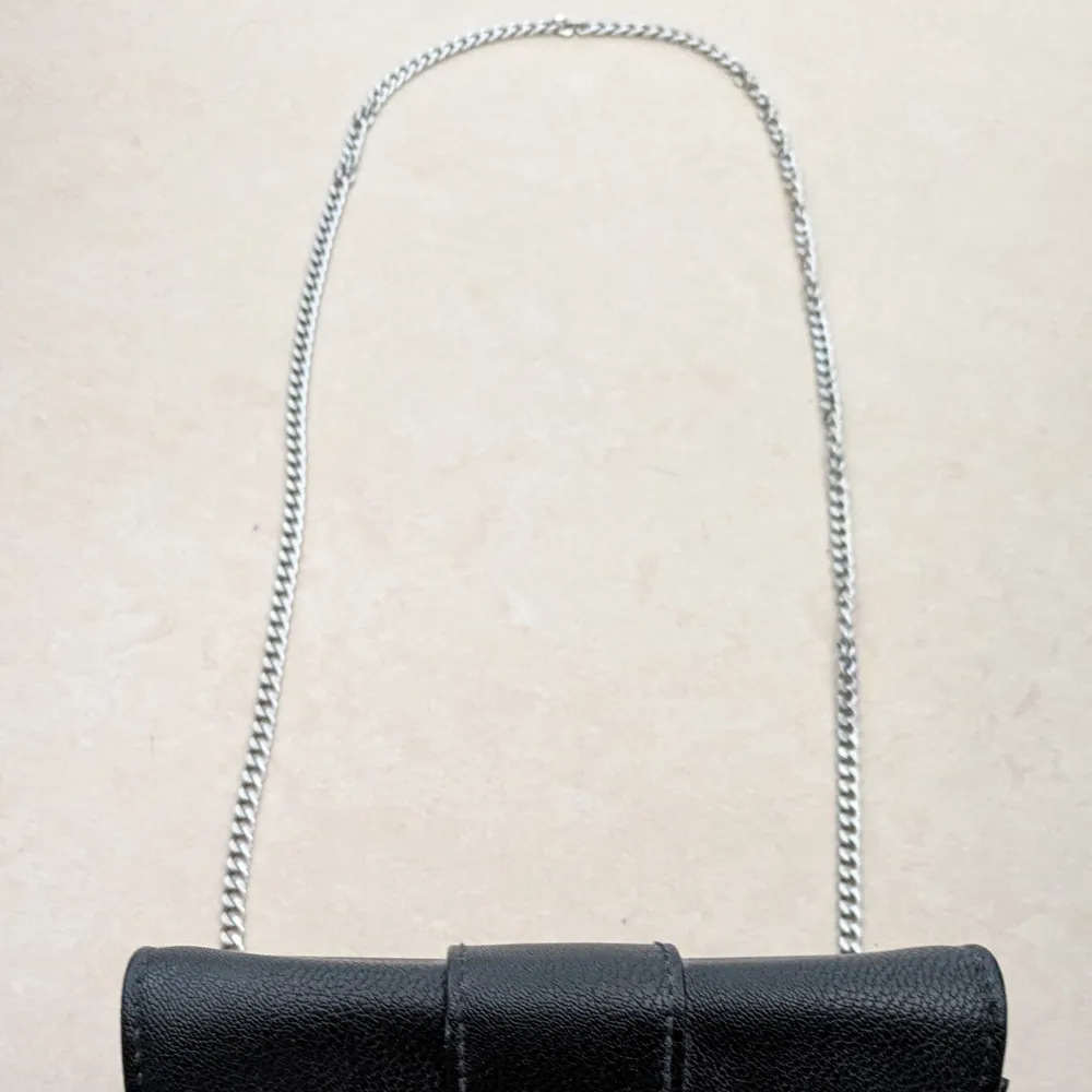 Elegant svart axelväska i syntetmaterial med en glittrig spänne framtill. Väskan har en silverfärgad kedja som axelrem, vilket ger en stilren look. Perfekt för att lyfta din outfit med en touch av glamour.. Laukut & Käsilaukut.
