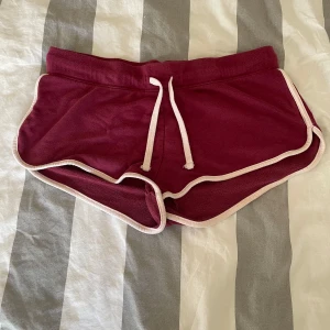 Vinröda shorts från H&M - Säljer ett par vinröda shorts från H&M med vita kanter och snörning i midjan. Perfekta för en avslappnad stil. De har en liten ficka bak och är gjorda i mjukt material.