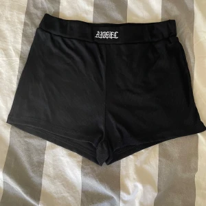 Svarta shorts med textdetalj - Snygga svarta shorts med en textdetalj 'ANGEL' i vitt på midjan. De är ribbade och har en bekväm passform, perfekta för en avslappnad stil.