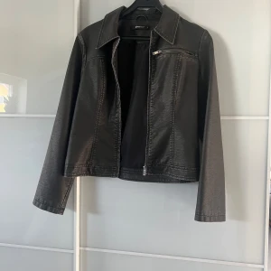 Svart skinnjacka från Gina Tricot - Aldrig använd! Snygg svart skinnjacka från Gina Tricot med dragkedja och klassisk krage. Jackan har en stilren design med långa ärmar och en liten bröstficka med dragkedja. Perfekt för en cool look!