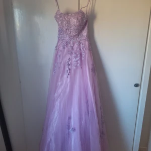 Ljuslila balklänning med spetsdetaljer - Elegant ljuslila balklänning med tunna axelband och vackra spetsdetaljer. Klänningen har en lång, flödande kjol och snörning i ryggen för en perfekt passform. Perfekt för speciella tillfällen.