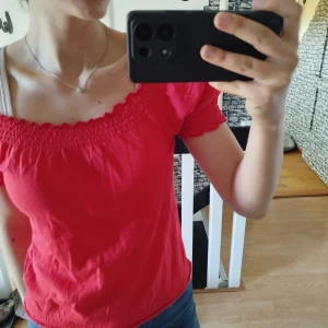 Röd offshoulder topp - Säljer en snygg röd offshoulder topp med korta ärmar. Toppen har en smockad detalj vid halsringningen och ärmarna, vilket ger en fin passform. Perfekt för sommarens varma dagar!