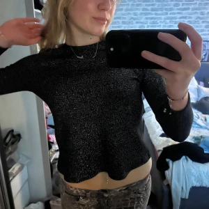 Svart glittrig topp med öppen rygg - Snygg svart glittrig topp med långärmad design och en stilfull öppen rygg. Perfekt för en kväll ute eller festliga tillfällen. Toppen har en elegant passform och ger en trendig look.