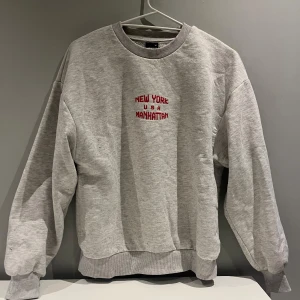 Grå sweatshirt från Gina Tricot - Säljer en grå sweatshirt från Gina Tricot med texten 'New York USA Manhattan' i rött på framsidan. Tröjan har en klassisk rund hals och långa ärmar. Tröjan är i fint skick🩶