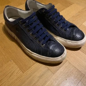 Blå sneakers från MLYN & HMLTN - Snygga blå sneakers med krokodilpräglat mönster och snörning. Perfekta för en stilren look med en touch av elegans. Skorna har en vit sula som kontrasterar snyggt mot det mörkblå materialet. köpta för 2399