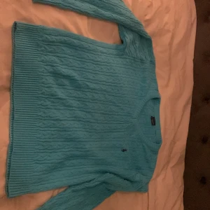 Turkos stickad tröja från Ralph Lauren - Säljer en snygg turkos stickad tröja från Polo Sport. Tröjan har en klassisk kabelstickad design med ribbade muddar och en liten broderad logga på bröstet. Perfekt för våren. Inga tecken på användning och extremt skönt material