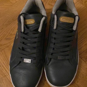 Gråa  sneakers från Lacoste - Snygga gråa sneakers från Lacoste med klassisk design. Skorna har snörning och en vit sula som kontrasterar mot det svarta skinnet. Perfekta för en stilren look.