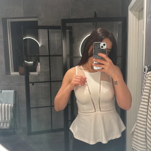 Vit ärmlös topp med meshdetaljer - Säljer en stilren vit ärmlös topp med transparenta meshdetaljer. Toppen har en peplumdesign som ger en fin siluett och en modern look. Perfekt för att klä upp en outfit!