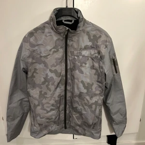 Camouflage vindjacka från Galvin Green - Säljer en sprillans ny och funktionell vindjacka från Galvin Green, helt oanvänd. Jackan har en dragkedja framtill och en praktisk ficka på ärmen. Perfekt för utomhusaktiviteter och håller dig skyddad mot vinden. Väldigt snygg till golftider också!!