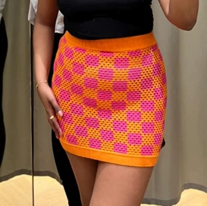 Rutig kjol i orange och rosa - Trendig kortkjol i ett färgglatt rutmönster med orange och rosa. Kjolen har en stickad struktur och en elastisk midja för en bekväm passform. Perfekt för att liva upp garderoben med lite färg!