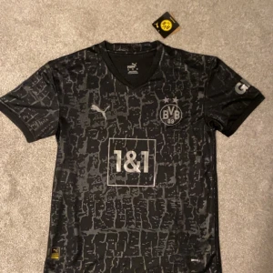 Svart BVB 09 fotbollströja från Puma - Snygg svart BVB 09 fotbollströja från Puma med ett unikt mönster. Tröjan har korta ärmar och klubbens logga på bröstet. Perfekt för fans som vill visa sitt stöd med stil. 🖤