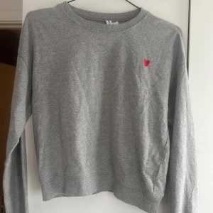 Grå sweatshirt från H&M Divided - Säljer en grå sweatshirt från H&M Divided med ett litet rött hjärta broderat på bröstet. Tröjan har en klassisk rund hals och långa ärmar. Perfekt för en avslappnad stil. Skicka ett meddelande vid intresse! 💌 materialet på tröjan är lite nopprigt