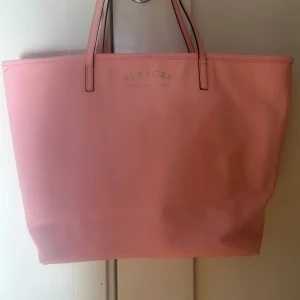 Rosa shoppingväska från H&M - Snygg rosa shoppingväska med texten 'New York' tryckt i guld på framsidan från H&M. Väskan har två handtag och är perfekt för att bära med sig allt man behöver under dagen.