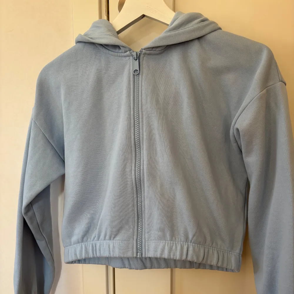 Säljer en ljusblå hoodie med dragkedja och huva. Hoodien har långa ärmar och en elastisk nederkant för en bekväm passform. Perfekt för en avslappnad stil.. Hupparit & Collegepaidat.