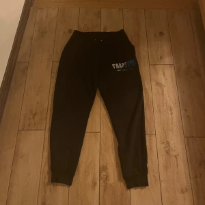 Svarta mjukisbyxor från Trapstar - Säljer ett par svarta mjukisbyxor från Trapstar med elastisk midja och muddar vid bensluten. Byxorna har en tryckt logga i blått och vitt på ena benet. Perfekta för en avslappnad stil.