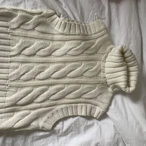 Vit stickad väst från H&M - Säljer en snygg vit stickad väst från H&M med ett klassiskt flätmönster. Västen har ribbade kanter vid hals och ärmslut, perfekt för lager-på-lager-stil. Passar utmärkt till både vardag och speciella tillfällen.