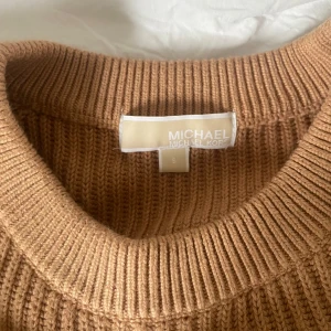 Beige stickad tröja från Michael Kors - Säljer en snygg beige stickad tröja från Michael Kors. Tröjan har en ribbad design och långa ärmar, perfekt för kyligare dagar. Den har en rund halsringning och en diskret logga i guld på framsidan. Perfekt för en stilren look!