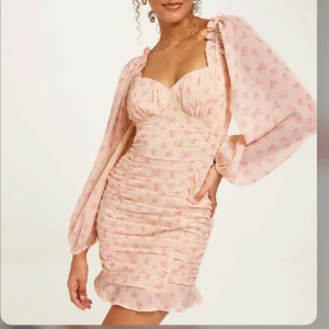 Ljusrosa/aprikos blommig klänning - Charmig ljusrosa/aprikos klänning med blommigt mönster och volangdetaljer. Klänningen har långa ärmar och en v-ringad design med rynkade detaljer som ger en smickrande passform.