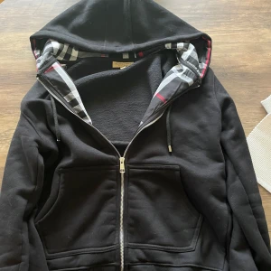 Svart huvtröja från Burberry - Säljer en stilren svart huvtröja från Burberry med dragkedja och klassiskt rutigt mönster i huvan. Tröjan har en broderad logga på bröstet och är perfekt för en avslappnad look. Den har långa ärmar och en mjuk insida för extra komfort.