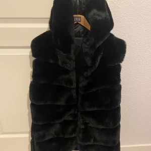 Svart pälsväst - Snygg svart pälsväst som ger en lyxig känsla. Perfekt för att hålla sig varm och stilren under kyliga dagar. Västen har en mjuk och fluffig textur.