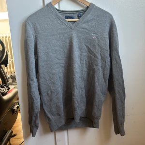 Grå tröja från Gant - Säljer en stilren grå tröja från Gant med v-ringning och broderad logga på bröstet. Tröjan har långa ärmar och är perfekt för en avslappnad men snygg look.