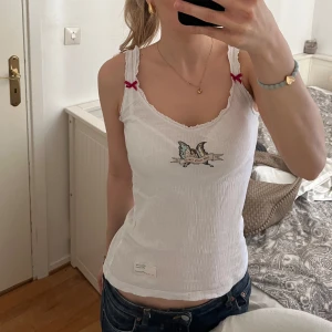 Vit  linnetopp med fjärilsbroderi - Säljer en söt vit ribbad linnetopp med ett broderat fjärilsmotiv på framsidan. Toppen har smala axelband med små röda rosetter och en fin vågig kant vid halsringningen. Perfekt för en avslappnad stil!