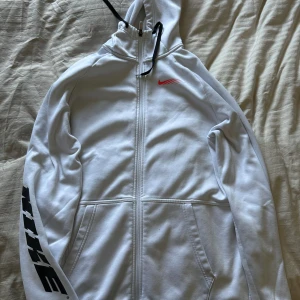 Vit hoodie med dragkedja från Nike - Säljer en vit hoodie med dragkedja från Nike. Den har en röd logga på bröstet och svart text på ärmen. Perfekt för en sportig look med sin bekväma passform och praktiska fickor.