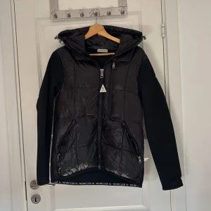  Moncler jacka - Snygg svart jacka från Moncler med huva och dragkedja. Jackan har långa ärmar och en quiltad design på framsidan. Moncler-logotypen finns på ärmen och insidan har en färgglad etikett med serietidningsmotiv.