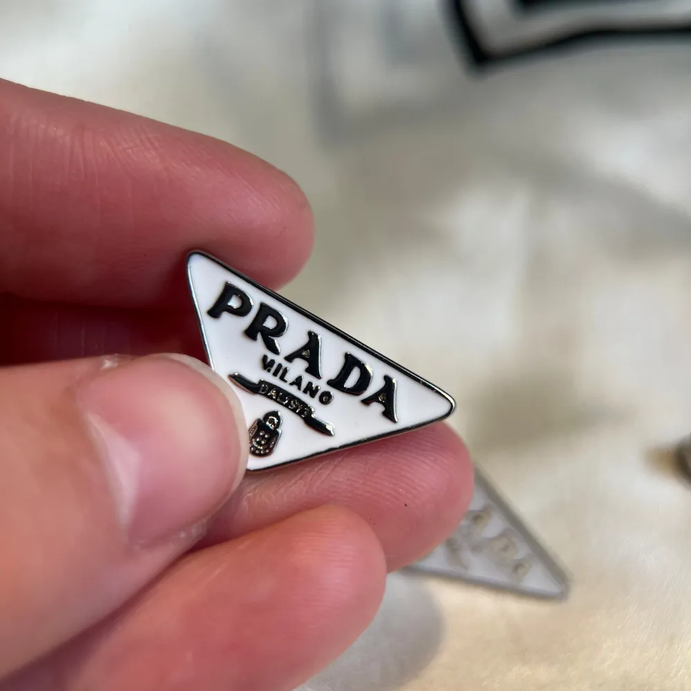 Snygga triangulära örhängen från Prada i silver och vit färg. De har en stilren design med Pradas logotyp tydligt synlig på framsidan. Perfekta för att ge en lyxig touch till din outfit.. Asusteet.
