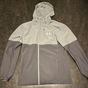 Grå vindjacka från Under Armour - Säljer en grå vindjacka från Under Armour med huva och dragkedja. Jackan har en stilren design med två nyanser av grått och en bröstficka med dragkedja. Perfekt för utomhusaktiviteter.