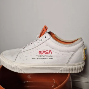 Vita sneakers med NASA-tryck från Vans  - Unika vita sneakers med NASA-tryck och detaljer i orange. Skorna har snörning och en robust sula med mönster för bra grepp. Perfekta för den som vill ha en rymdinspirerad stil.