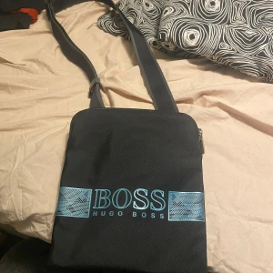 Svart axelväska från Hugo Boss - Snygg svart axelväska från Hugo Boss med justerbar rem och dragkedja upptill. Väskan har en stilren design med märkets logotyp i blått framtill. Perfekt för att bära dina dagliga nödvändigheter med stil.