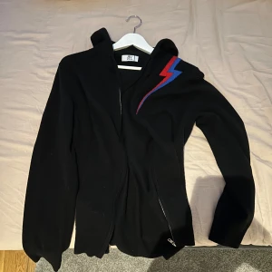 Svart cashmir x merinowool zip hoodie från BVALDI - Snygg svart hoodie från BV x AQC med en cool blixtdetalj i rött och blått på axeln. Tillverkad i en lyxig blandning av kashmir och merinoull, perfekt för att hålla dig varm och stilren. Dragkedja framtill och huva för extra komfort. Storlek medium. Lite kort på mig som är 187. Tyget är så sjukt skönt och tunnt så perfekt nu till våren. Enbart använd 1 gång. Nypris 1300kr säljer för 999