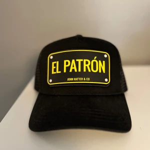 Svart keps från John Hatter & Co - Snygg svart keps med texten 'El Patrón' i gult på framsidan. Kepsen har ett nätmaterial på baksidan för bättre ventilation och en justerbar rem för perfekt passform. Perfekt för en cool och avslappnad stil. Använd cirka 4 gånger. Anledningen till att jag säljer den är för att jag inte passar i den. Vid några frågor har av er 😊