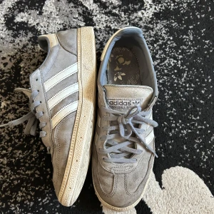 Grå Adidas spezial  - Säljer ett par grå Adidas sneakers i mocka med de klassiska tre vita ränderna på sidan. Skorna har snörning och en vit sula. Perfekta för en avslappnad stil.